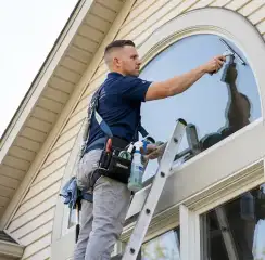 Professional window cleaner on a ladder using a squeegee to clean an exterior arched window, El Paso, TX.